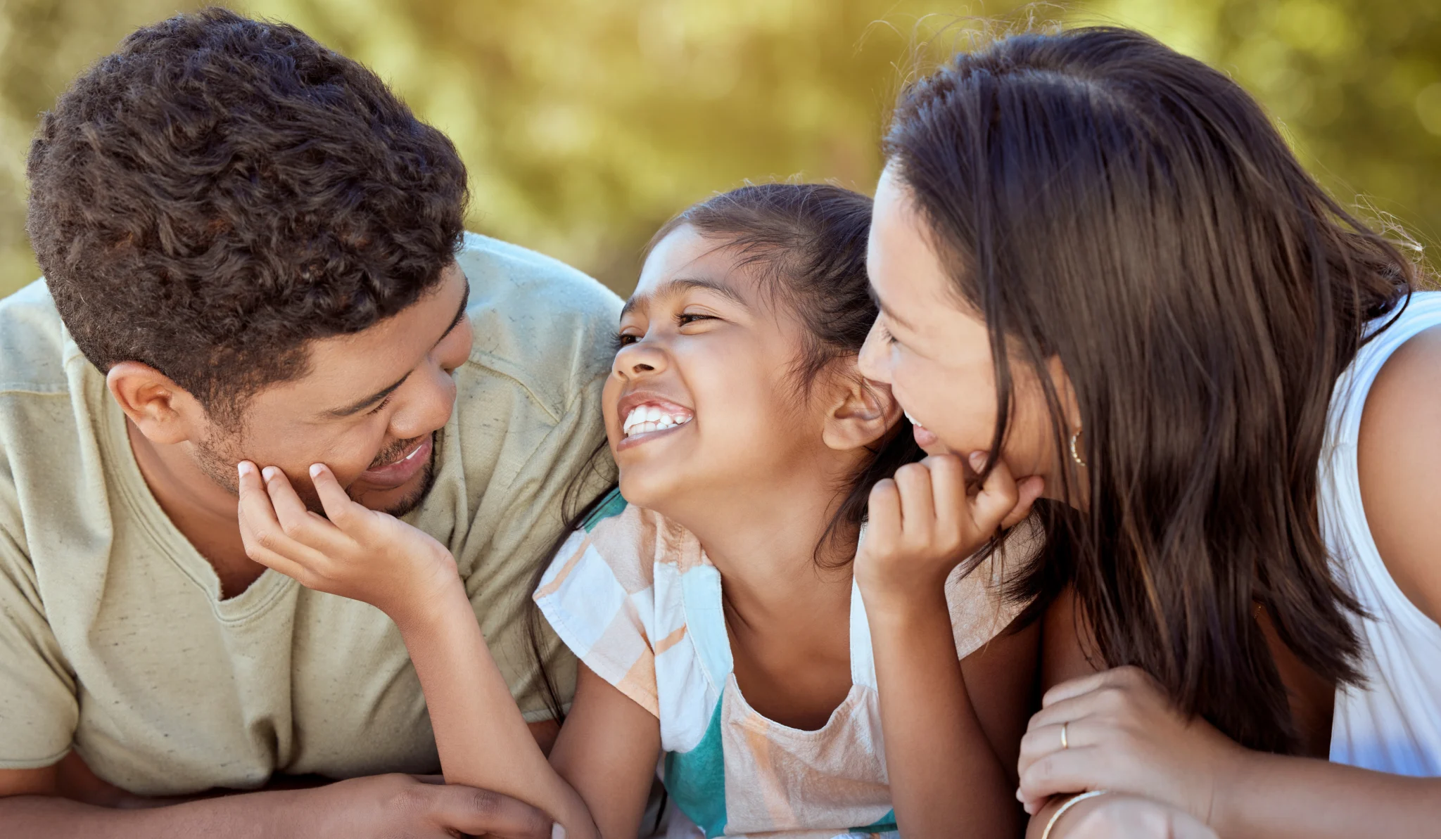 Pai, mãe e filha trocam sorrisos próximos, curtindo um momento de lazer em família ao ar livre.